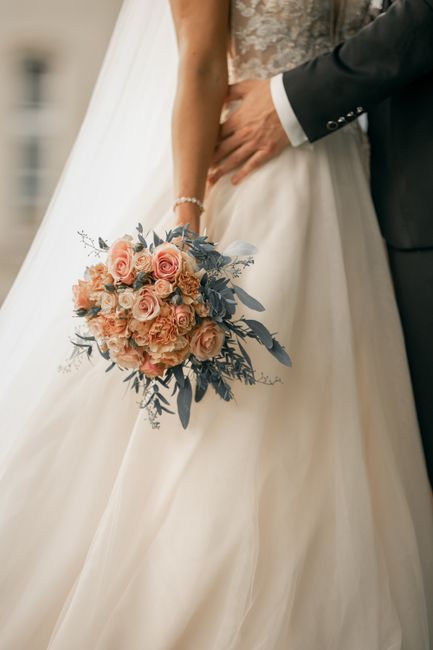A bride in a white lace wedding dress holding a large bouquet of fresh roses in soft peach and dusty pink tones, accented with blue-grey eucalyptus and foliage. The bouquet is arranged in a rounded, l