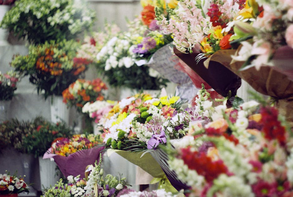 A display of various floral arrangements and bouquets featuring an array of fresh flowers in vibrant colors such as pink, yellow, white, purple, orange, and red. The flowers include lilies, carnations