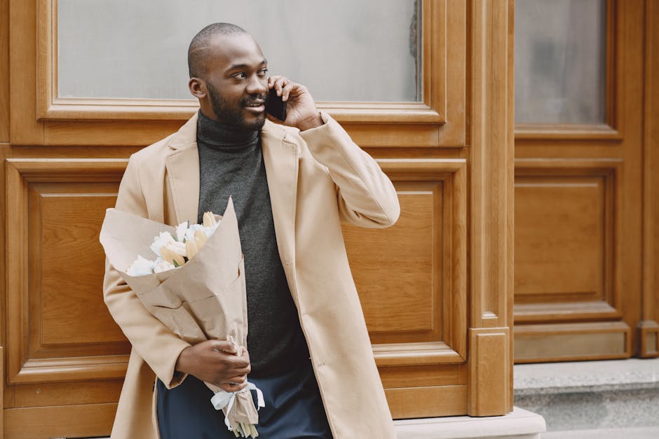 A man with a dark complexion, short hair, and a beard is standing outdoors against a wooden-paneled wall, holding a bouquet of fresh white flowers wrapped in brown paper. He is dressed in a beige coat