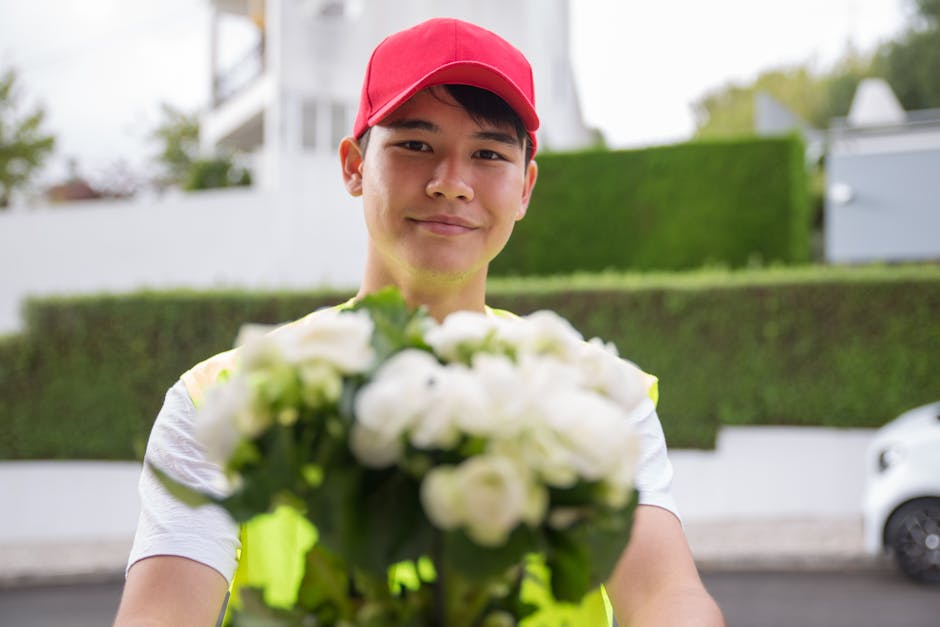A young delivery person wearing a bright yellow high-visibility vest and a red baseball cap is holding out a floral bouquet towards the camera, standing outdoors in a residential area with white build
