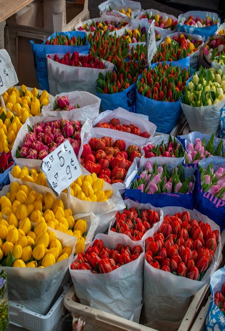 An array of tulip bouquets arranged in large paper wraps at a flower market display, showcasing vibrant colors including yellow, pink, red, orange, and green, with some bouquets having visible stems a