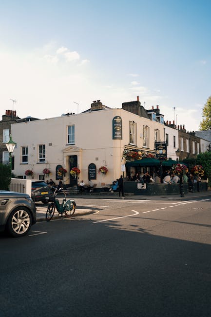 A street scene featuring a white, two-story building with traditional pub-style signage and green awnings, adorned with hanging flower baskets and window boxes filled with vibrant red, pink, and purpl