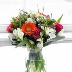This elegant arrangement captures the lively essence of Chiswick, echoing the gentle beauty of morning markets and riverside strolls. The bouquet features clusters of orange and peach gerberas, their petals radiating outward in a soft halo, with deep brown hearts reminiscent of the chestnut trees in Gunnersbury Park. Between these, fuchsia-mauve roses bloom in luxurious tiers, their dense structure imparting drama, while pearl-white alstroemeria and petite spray roses float among the larger forms like wisps of morning mist. Subtle dashes of rose-pink blossoms heighten the sense of abundance, making the composition feel both curated and spontaneous. Large, smooth aspidistra leaves, deep and lustrous, are gathered into a protective ring around the base, supporting the bouquet's full, rounded appearance. Threads of pointed green leaves and delicate sprigs bring natural rhythm and movement, as if plucked fresh from a carefully tended Chiswick garden. Presented in a clear vase that reveals sturdy, vital stems, the arrangement feels alive, accentuated by the quietly sparkling white background that mirrors the light-filled interiors of local studios. A bouquet for joyful occasions, new beginnings, or thoughtful tokens of appreciation, this display speaks of care, artistry, and the enduring charm of Chiswick's neighborhoods.
